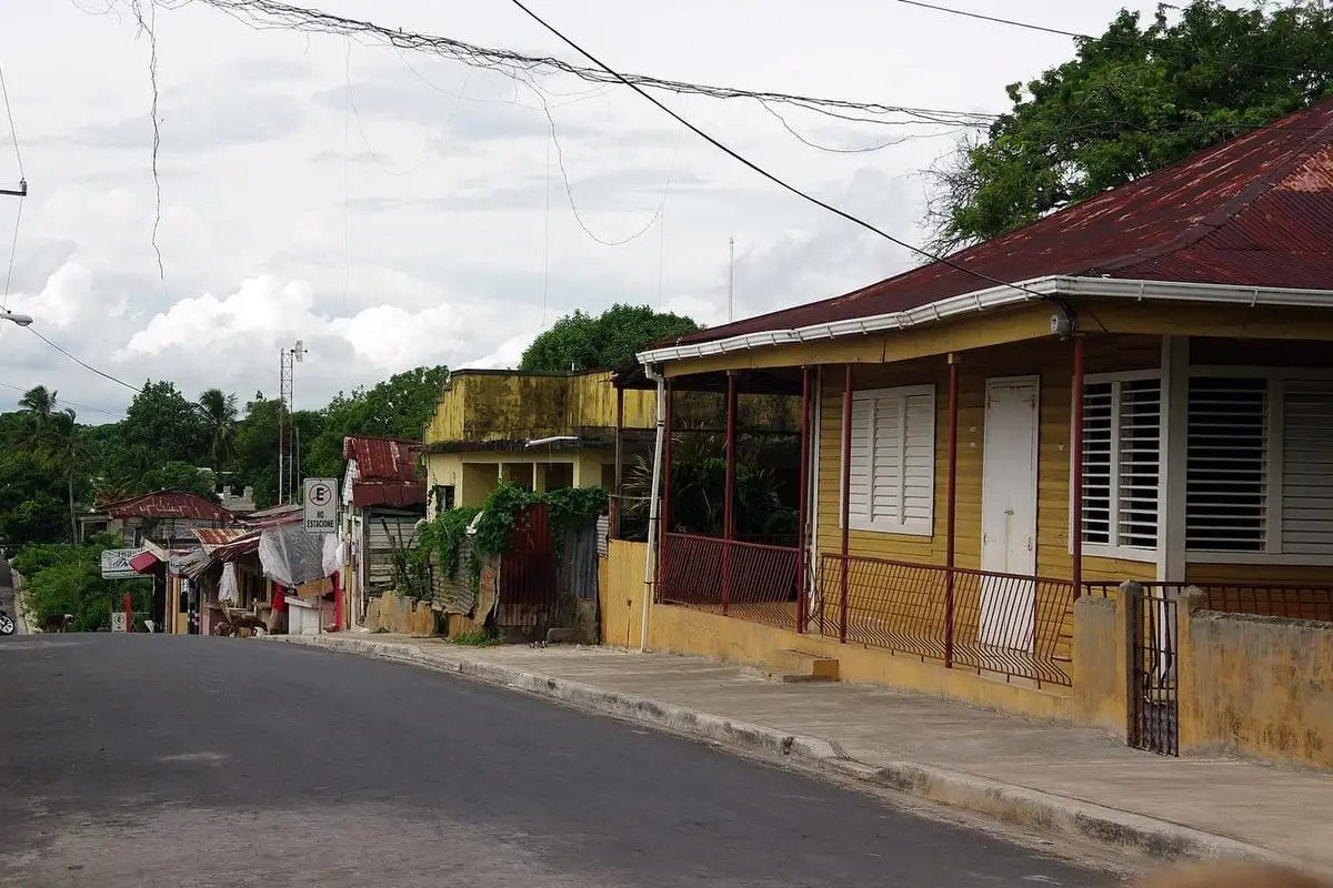 Calle en bajada en Sánchez, con una casa de madera amarilla de techo de zinc rojo y galería con barandas, bajo un cielo nublado