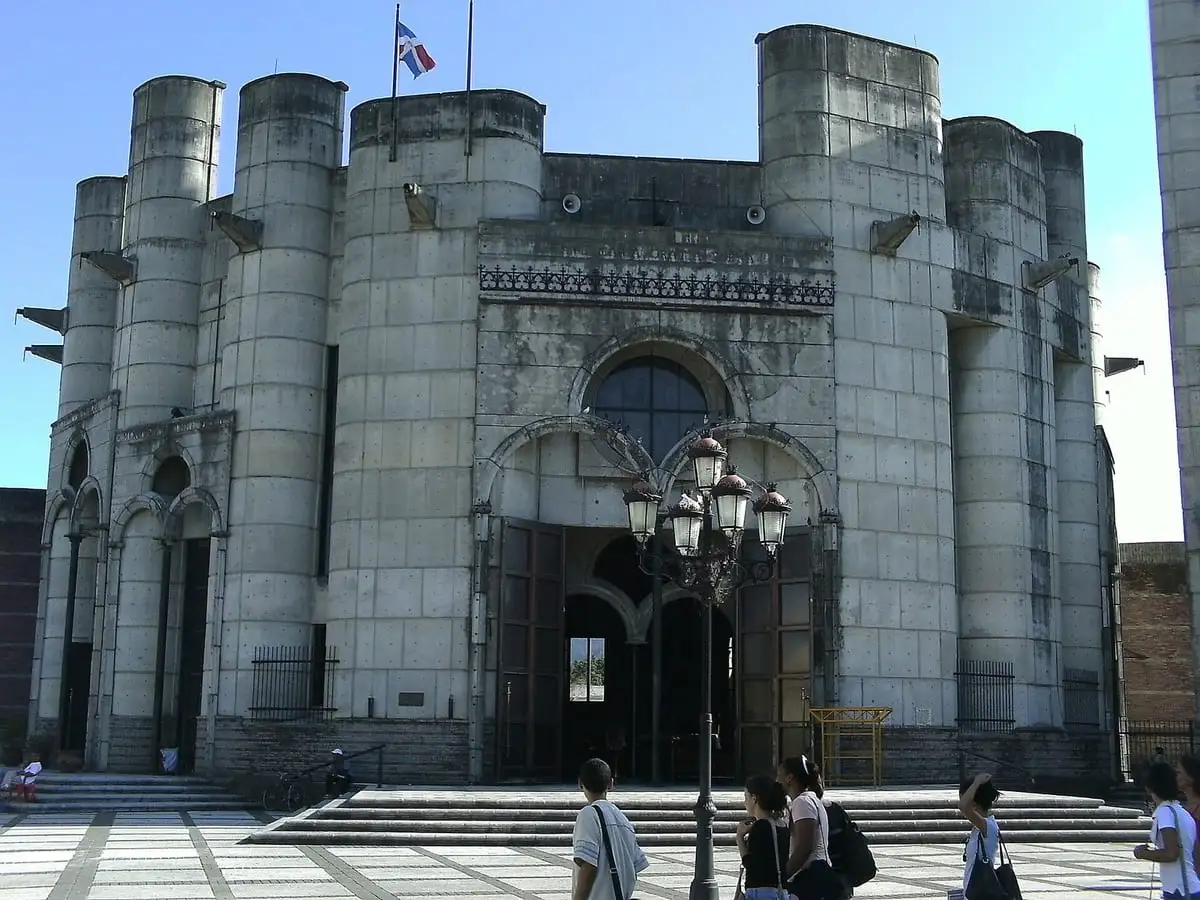 Fachada de hormigón gris de la catedral de La Vega, con torres cilíndricas, un gran ventanal en arco y la bandera dominicana arriba