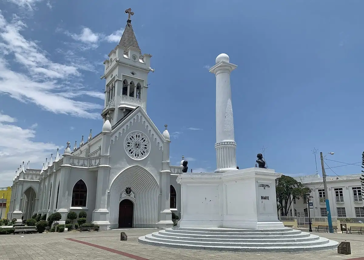 Catedral San Pedro Apóstol, blanca y de torre en punta, junto a una columna blanca en la plaza de San Pedro de Macorís