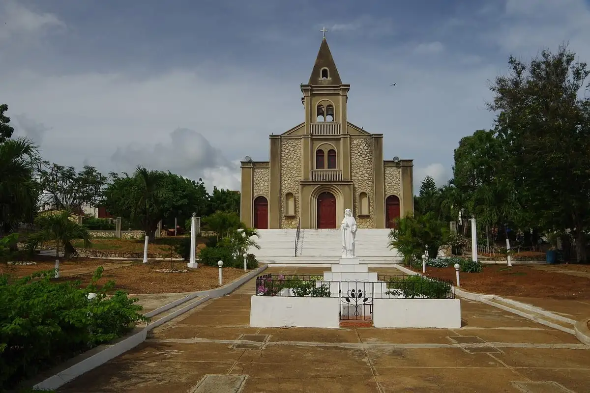 Iglesia Santa Rosa de La Romana, de fachada color arena y torre en punta, al final de un paseo con una estatua blanca