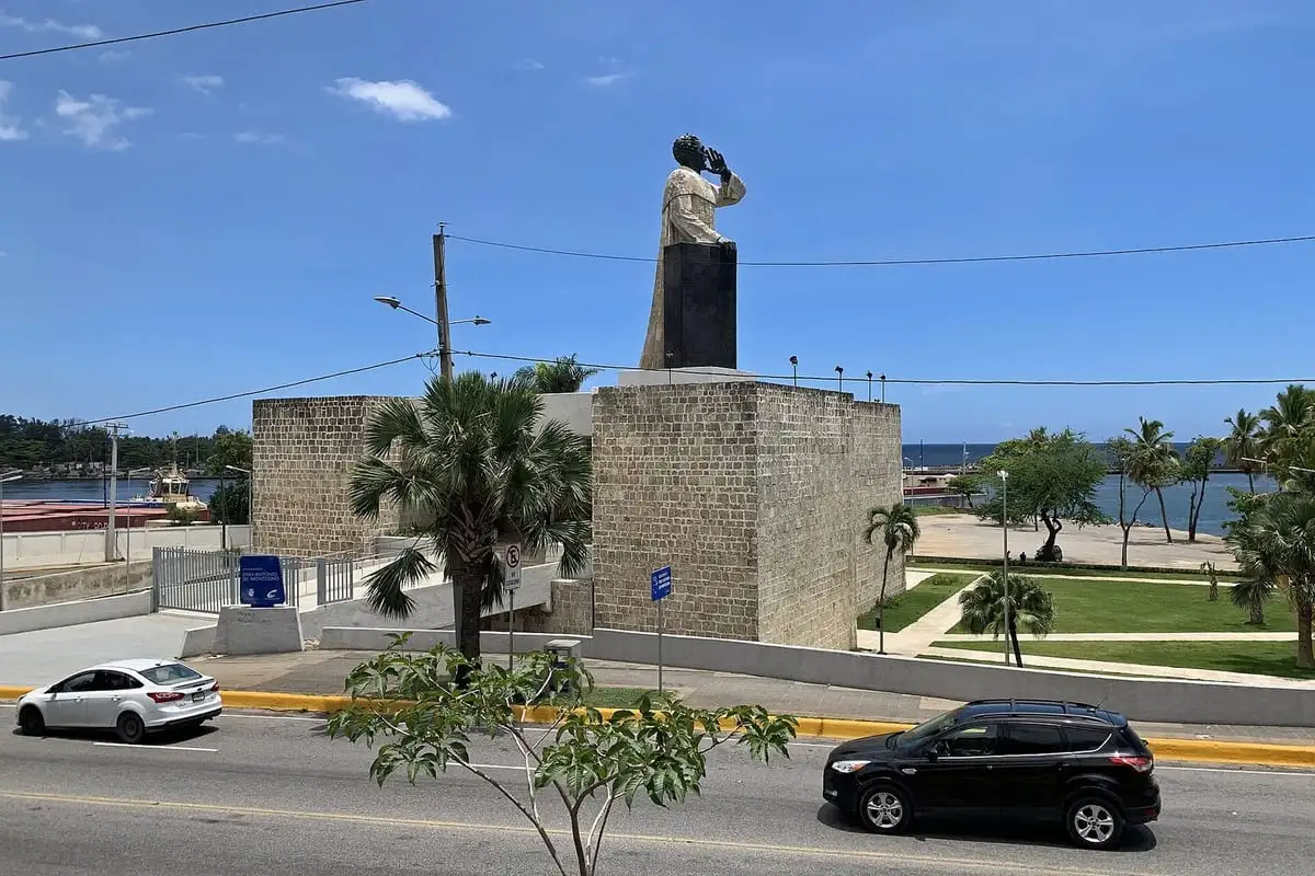 Monumento a Fray Antón de Montesinos, una figura sobre un gran pedestal de piedra frente al mar, con carros pasando por el Malecón