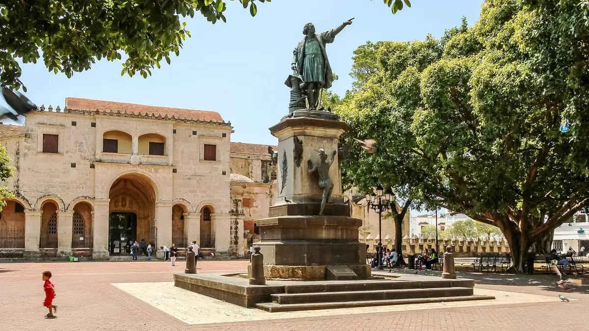 Estatua de Cristóbal Colón señalando con el brazo en el Parque Colón, con la Catedral de piedra al fondo y árboles grandes a los lados