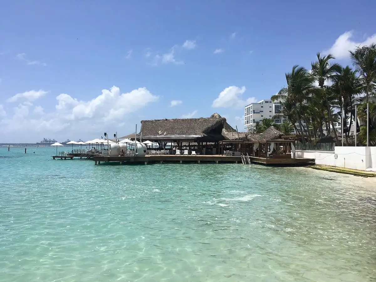 Agua turquesa y poco profunda en la playa de Boca Chica, con un restaurante de techo de cana sobre el mar y palmeras detrás